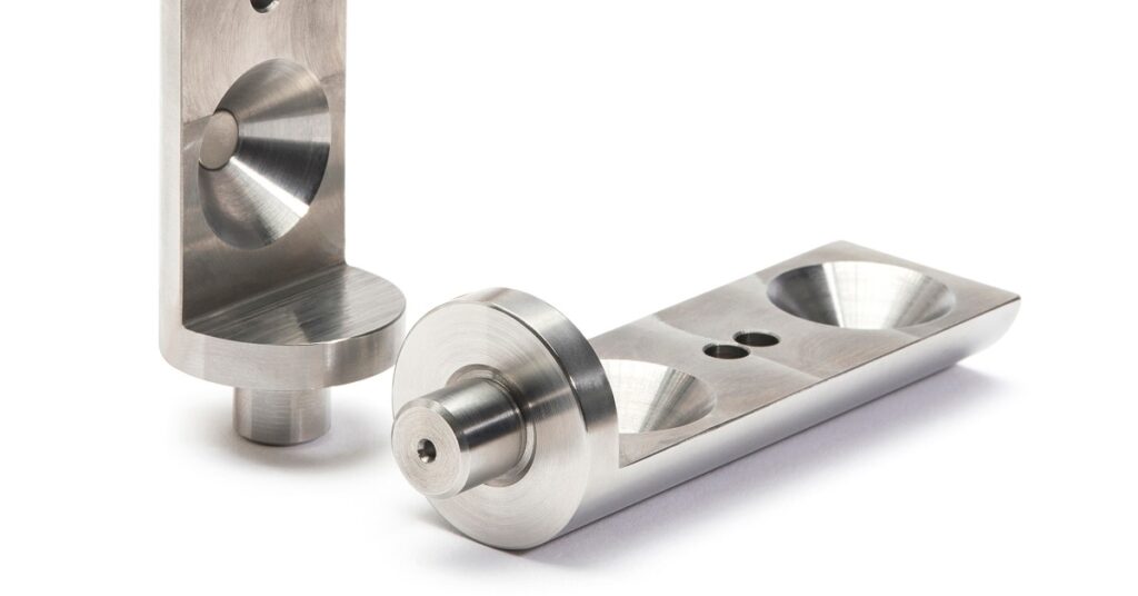 Three silver machined metal parts with round recesses, angled edges, small holes, and brushed surfaces.