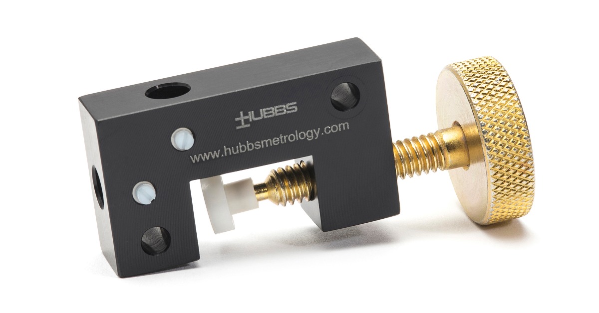 A black metal measurement tool with a brass threaded knob, white pins, round holes, and printed website text.